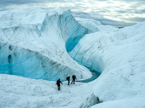 Group of hikers exploring a stunning glacier with deep blue ice crevasses under a cloudy sky. Greenland icecap