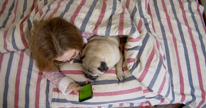 Beauty teen girl and her cute pug dog lying in the bed, watching, looking at smartphone. Green screen. Friendship with pet concept. Together forever, inseparable friends
