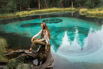 A beautiful girl sits on a very beautiful and turquoise geyser lake in Russia, in the Altai Republic. National Reserve. Aqua Menthe color. Mixed forest with pines and cedars