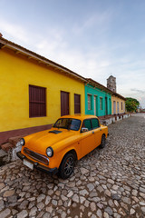 Fototapeta premium View of an Old Classic Taxi Car in the streets of a small Cuban Town with Church in the Background during a vibrant sunny sunrise. Trinidad, Cuba.