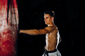 Fototapeta na wymiar Attractive shirtless male boxer practicing on a punching bag indoors
