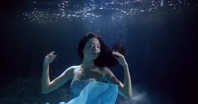 A portrait of a dark-haired girl with rhinestones on her face, who is underwater, the delicate fabric of her dress develops, she makes movements with her hands and emerges from the water.
