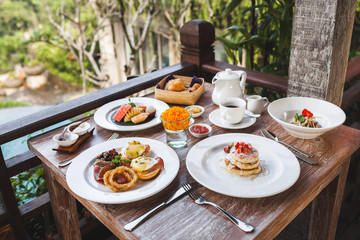 Traditional breakfast with eggs Benedict and strawberry pancakes on wooden table on terrace. Fruit plate and cup of coffee