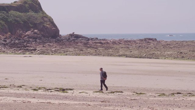 A daylight wide shot of a man carrying a knapsack on his back and walking on the sandy grounds of a rocky shoreline.