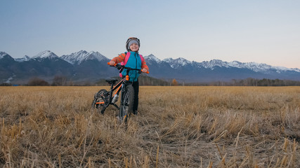 Fototapeta premium One caucasian children walk with bike in wheat field. Little girl walking black orange cycle on background of beautiful snowy mountains. Biker stand with backpack and helmet. Mountain bike hardtail.