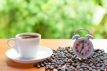 Alarm clock is seven o'clock in the morning.Coffee cup with the soft green of the forests and Coffee beans on the table. People drink coffee before work or travel.