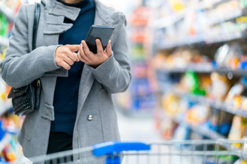 closeup asian female hand hold smartphone checking price compare in supermarket store with trolley...