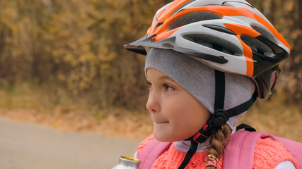Fototapeta na wymiar Kid drink water from an aluminum flask bottle. One caucasian children rides bike road in autumn park. Little girl riding black orange mtb cycle in forest. Biker motion ride with backpack and helmet.