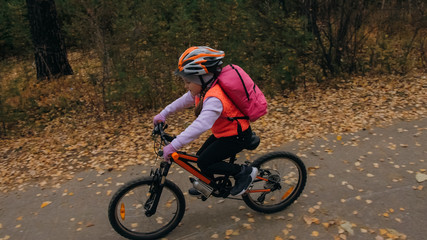 Fototapeta premium One caucasian children rides bike road in autumn park. Little girl riding black orange cycle in forest. Kid goes do bicycle sports. Biker motion ride with backpack and helmet. Mountain bike hardtail.