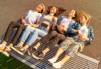 top view of four smiling teenagers lying with their legs on a bench in square at sunset