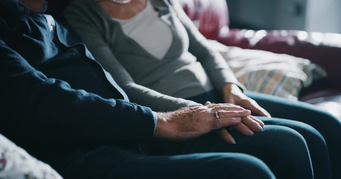 Close up of a lovely senior couple is holding a hand of each other with affection as sign of forever love and respect to each other in on sofa in living room at home.