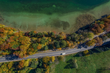 Aerial view of road with a car in autumn forest next to a touquise colored lake.