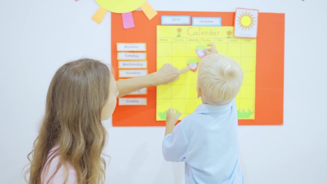 Preschool boy adhering a heart symbol on calendar board with his teacher in the classroom. Shot in 4k resolution