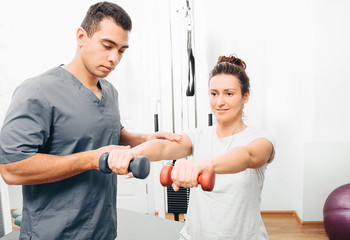 Physiotherapist working with male patient in clinic. Raises dumbbells, strengthening arm muscles and developing joints