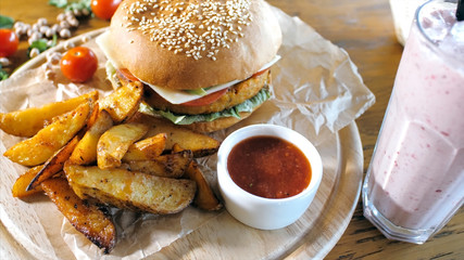 Crispy rustic potatoes with hamburger and red sauce on wooden board next to pink smoothie, close-up. Fast food concept with no people.