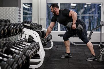 strong young bearded shouting man pull dumbbell in one hand standing in slope in sport gym