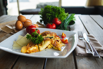 Traditional omelet with vegetables, spinach, tomatoes and herbs on a wooden table in a restaurant. Classic european breakfast. Fried eggs with milk and cheese. Soft focus.