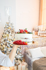 Cake decoration with lingonberry berries, blueberries and cotton. Women's hands in a frame in a bright kitchen. Christmas treat for the holiday. Lights of garlands.
