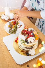 Cake decoration with lingonberry berries, blueberries and cotton. Women's hands in a frame in a bright kitchen. Christmas treat for the holiday. Lights of garlands.