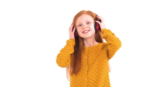 Portrait shot of a little charming girl with long red hair taking on wireless headphones. Cheerful girl starts dancing. True emotions concept. Isolated, on white background