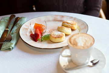 Cheesecakes, baked polpettine di ricotta on a plate with jam and strawberries. On the table is a cup of coffee, a glass of orange juice, a fork and a knife in hand