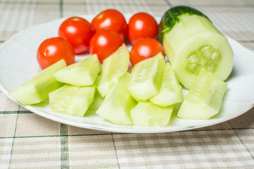 Small red tomatoes and sliced cucumber in plate on kitchen table for snack.