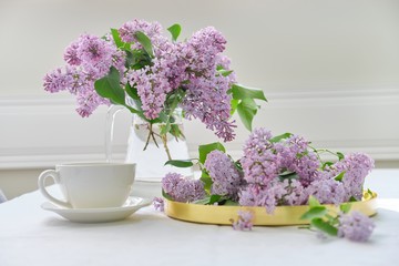Spring bouquet of lilac flowers in glass jug on table, cup of tea