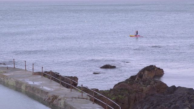 A daylight long shot of two tourists each paddling their boat in the ocean water near the rocky shoreline as seen from a distance.