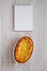 Homemade Cheddar Corn Pudding Casserole, blank notepad on a white wooden surface, top view. Flat lay, top view, from above. Copy space.