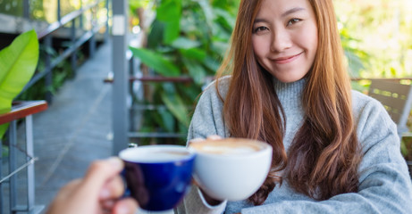 Close up image of a beautiful asian woman clinking coffee mugs with friend in cafe