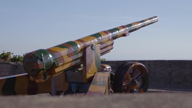 A daylight closeup shot of an artillery gun painted with a camouflage pattern and on display on top of a fort overlooking the sky.