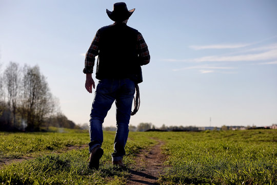A man cowboy hat and a loso in the field. American farmer in a field wearing a jeans hat and with a loso. A man is walking across the field silhouette