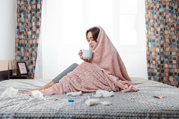 The concept of diseases and seasonal colds. A young brunette woman is sitting on the bed, wrapped...