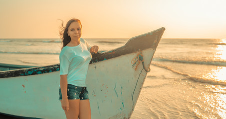A young woman stands near the boat by the sea against the sunset.