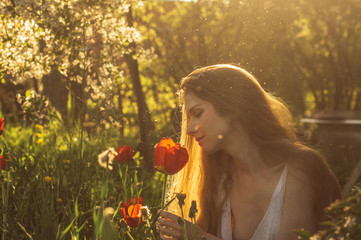 Naklejka premium Girl in white dress smelling tulip in sunset among fluff, dandelions and cherry flowers