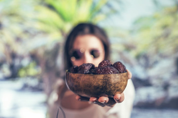 Woman in sunglasses holds out royal dates fruit in a bowl of coconut in a palm tree grove