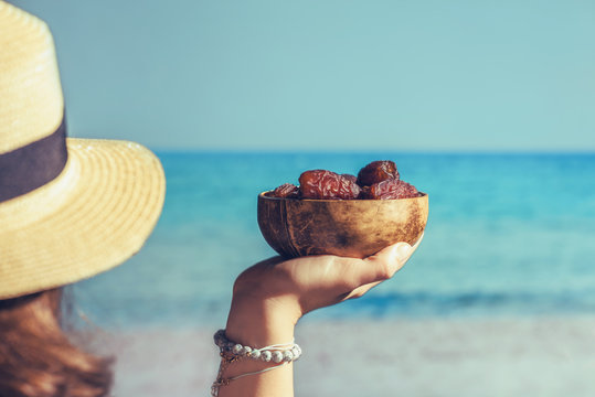 Woman In A Straw Hat Holding In A Hand Royal Dates Fruit In A Bowl Of Coconut On A Beach With Sea View.