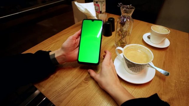 Young female hands using green screen smartphone resting in cafe drinking coffee with friends. Online business communication. Chromakey mock-up. Blank touchscreen. Social networking. Chatting. 