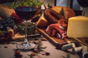 Table with gourmet ingredients, glass of wine, variety of cheeses, cold meats, grapes, raspberry, blackberry and artisan bread on a rustic background