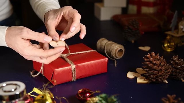 Close up on a women wrapping gifts for Christmas in red craft paper. The gift is decorated with strings and heart-shaped wood decorations.