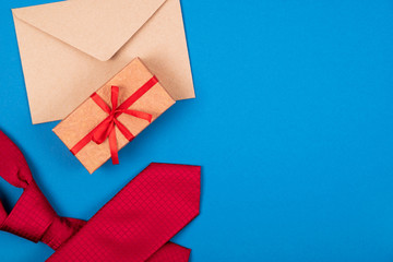 Naklejka premium Composition of red neck tie, gift box with bow and brown craft paper envelope on blue cyan background.