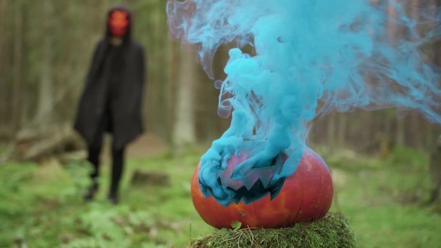 Person goes in mask and coak in the forest on the halloween with smoky pumpkin