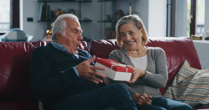 A lovely senior wife is making a surprise gift to her husband as sign of love for their anniversary in living rom at home.