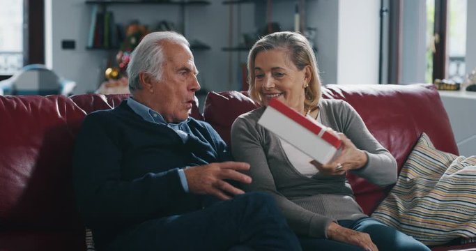 A lovely senior wife is making a surprise gift to her husband as sign of love for their anniversary in living rom at home.