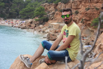 European caucasian boy in summer outfit on vacation in September in ibiza sitting on a cliff above the balearic sea