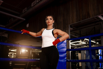 portrait of beautiful fit and sportive caucasian woman in white top and dark braided hair stand posing in ring, wearing red bandages on hands, ater fighting, boxing
