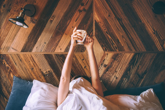 Lazy female lying under the white blanket on the  linen bed and holding the fresh coffee cup in the early morning. Lazy day off and "coffee in bed"concept image.