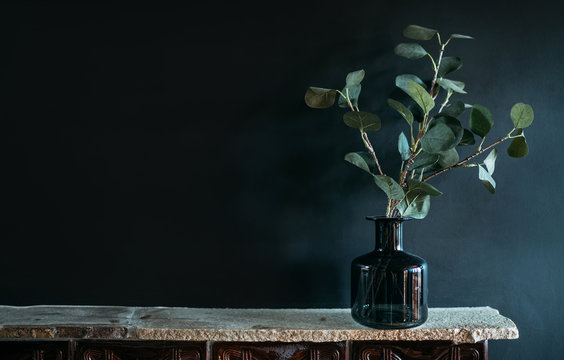 Green tree Branch putted into black glass vase on the natural stone mantel shelf on the black color wall background lit with side window light. Cozy home decor elements concept image.