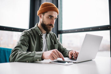 Handsome bearded man using laptop computer, sitting in office. Portrait of young pensive copywriter...