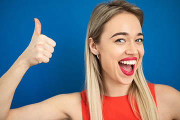 Happy emotional thumbs up beautiful blonde woman. Big adorable teeth smile. Red lips. Blue background